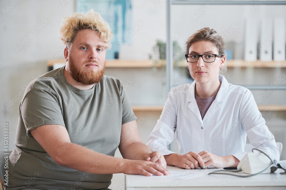 Portrait of young overweight man sitting at the table with his nutritionist and they looking at camera together at office Portrait of young overweight man sitting at the table with his nutritionist and they looking at camera together at office