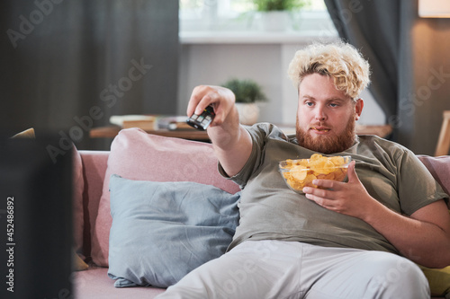 Obraz na plátně Lazy overweight man sitting on sofa with chips and using remote control to watch
