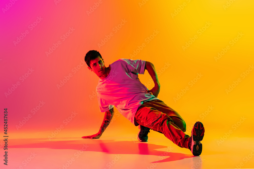 Solo performance. Young man, break dancing dancer ptacticing in casual clothes isolated over gradient pink yellow background at dance hall in neon light. Solo performance. Young man, break dancing dancer ptacticing in casual clothes isolated over gradient pink yellow background at dance hall in neon light.