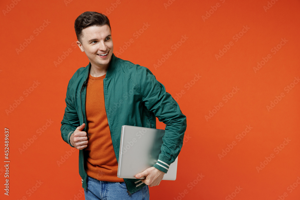 Smiling happy charming fun young brunet man 20s wears red t-shirt green jacket hold use work on laptop pc computer under his hand look back behind isolated on plain orange background studio portrait. Smiling happy charming fun young brunet man 20s wears red t-shirt green jacket hold use work on laptop pc computer under his hand look back behind isolated on plain orange background studio portrait.