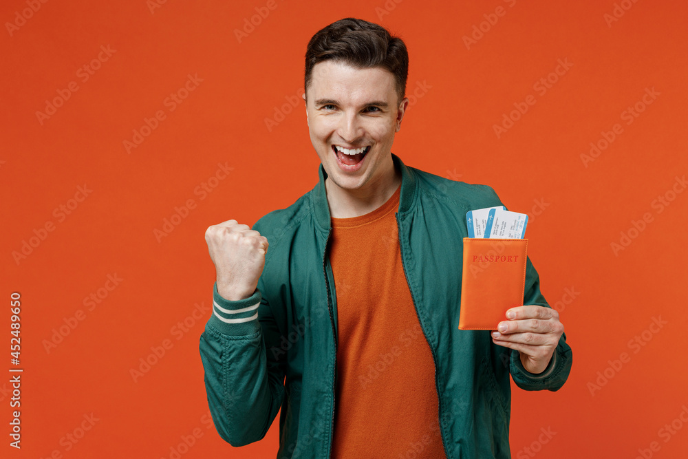 Traveler tourist excited overjoyed young brunet man 20s wear red t-shirt green jacket hold passport boarding tickets doing winner gesture celebrate isolated on plain orange background studio portrait Traveler tourist excited overjoyed young brunet man 20s wear red t-shirt green jacket hold passport boarding tickets doing winner gesture celebrate isolated on plain orange background studio portrait