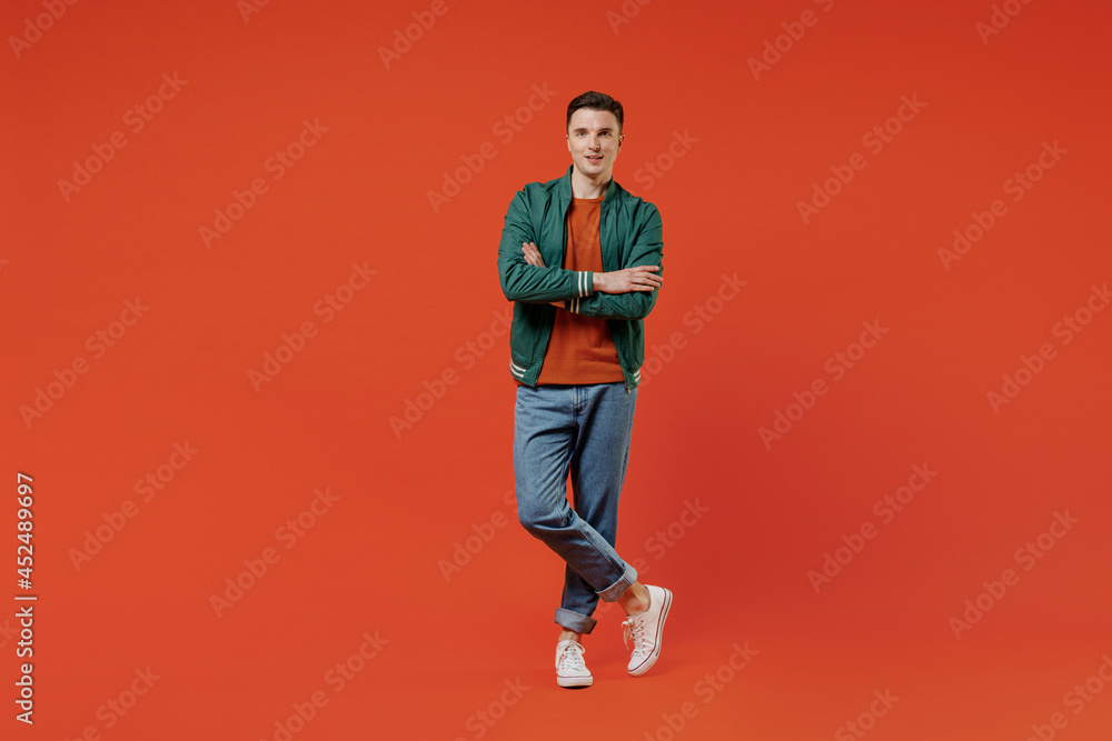 Full size body length smiling vivid happy young brunet man 20s wear red t-shirt green jacket hold hands crossed isolated on plain orange background studio portrait. People emotions lifestyle concept. Full size body length smiling vivid happy young brunet man 20s wear red t-shirt green jacket hold hands crossed isolated on plain orange background studio portrait. People emotions lifestyle concept.