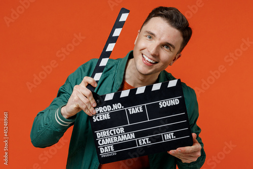 Smiling cheerful cool vivid happy young brunet man 20s wear red t-shirt green jacket holding hiding behind classic black film making clapperboard isolated on plain orange background studio portrait