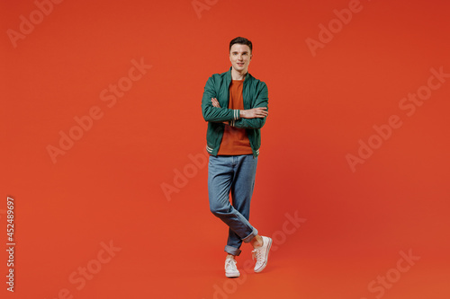 Full size body length smiling vivid happy young brunet man 20s wear red t-shirt green jacket hold hands crossed isolated on plain orange background studio portrait. People emotions lifestyle concept.