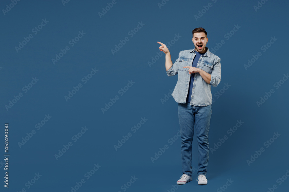 Full size length body beautiful happy young brunet man 20s wears denim jacket pointing on workspace area copy space mock up isolated on dark blue background studio portrait. People emotions concept