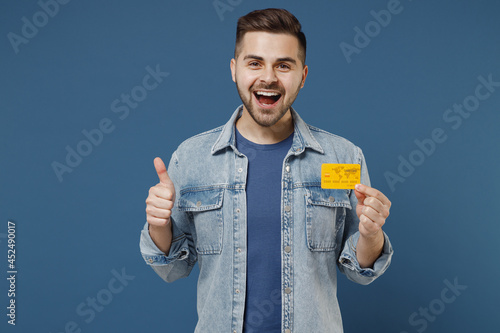 Quadro su tela Satisfied happy excited young brunet man 20s wears denim jacket hold in hand credit bank card show thumb up gesture isolated on dark blue background studio portrait