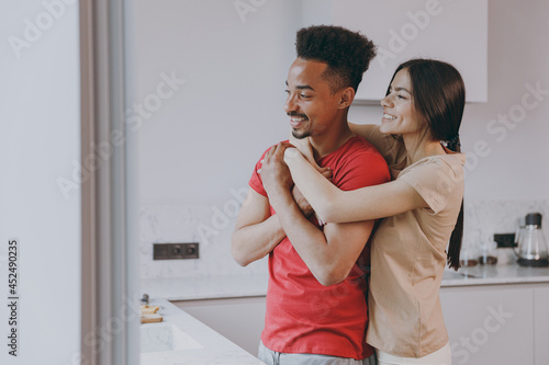 Young lovely cheerful married couple two woman american african man 20s wear casual t-shirt clothes wife hugging husband in light kitchen at home together Healthy diet lifestyle relationship concept photo