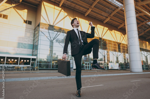 Bottom view full size young traveler brunet businessman man in black dinner suit stand outside at international airport terminal with briefcase diplomat do winner gesture Air flight business concept.