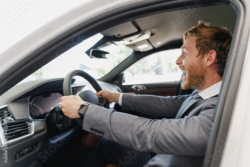 Side view excited man customer buyer client in classic grey suit driving car hold wheel choose auto want buy new automobile in showroom vehicle salon dealership store motor show indoor Sale concept. © ViDi Studio