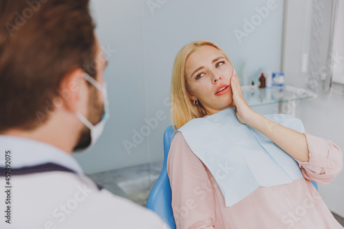 Young ill woman 20s look at dentist sit at office chair indoor light cabinet hold cheeck visiting stomatologist with toothache painful problem wait for cure Healthcare oral enamel caries treatment