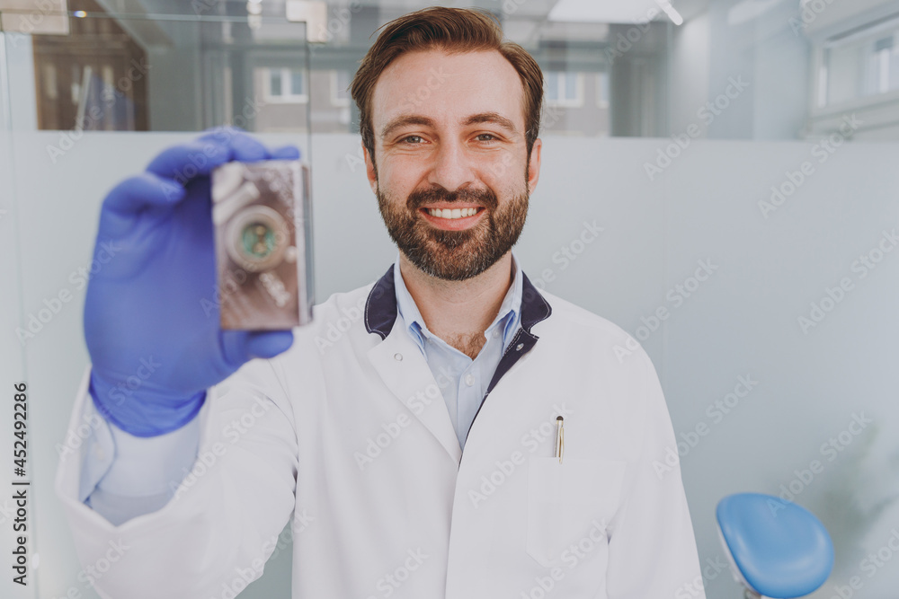 Young caucasian confident professional positive smiling happy dentist doctor man 30s holding dental implant at office indoor light modern cabinet near stomatologist Healthcare oral enamel treatment. Young caucasian confident professional positive smiling happy dentist doctor man 30s holding dental implant at office indoor light modern cabinet near stomatologist Healthcare oral enamel treatment.