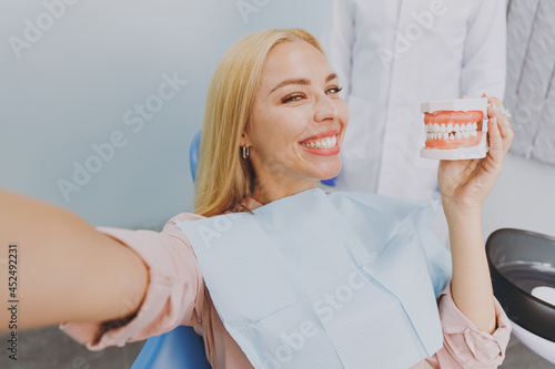Close up young cheerful woman do selfie shot pov on mobile phone hold artificial jaw sit at dental office chair indoor cabinet wait stomatologist for oral procedure. Healthcare orthodontal treatment photo