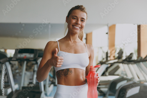 Young fun strong skinny sporty athletic sportswoman woman wear white sportswear warm up train near treadmill drink water hold pink bottle show thumb up in gym indoor Workout sport motivation concept. photo