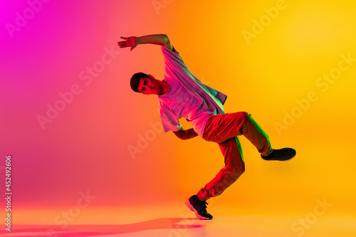 Portrait of young stylish man, break dancing dancer training in casual clothes isolated over gradient pink yellow background at dance hall in neon light.