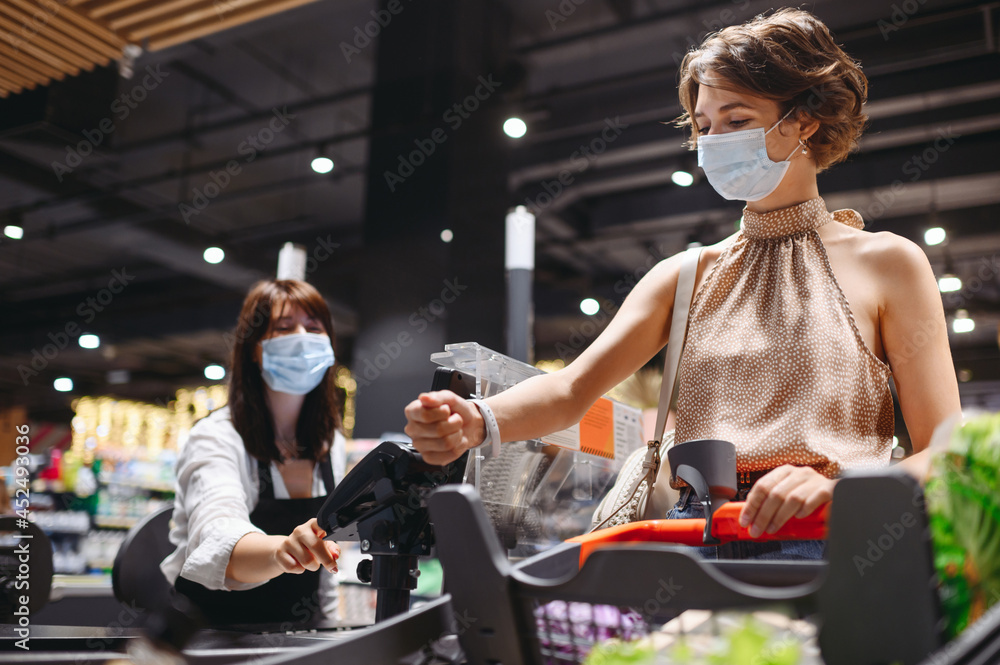Obraz premium Young caucasian woman 20s in casual clothes mask shopping at supermaket with grocery cart put smart watch to modern bank payment terminal process acquire payments near cashier checkout inside store.