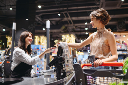 Tableau sur toile Young smiling woman wear casual clothes mask shopping at supermaket stand at store checkout giving credit card to cashier pay inside hypermarket