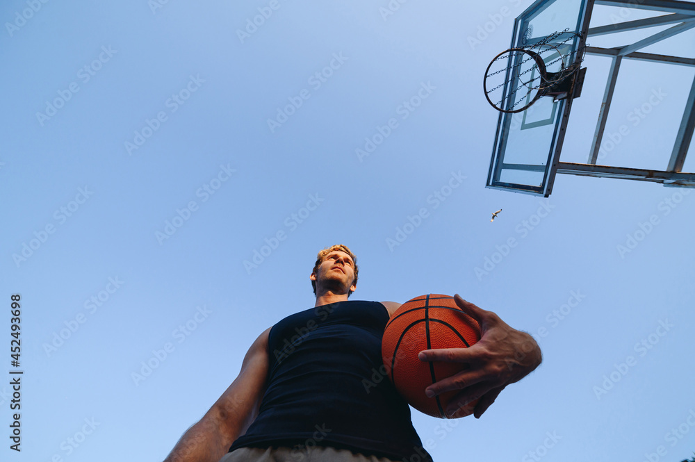 Bottom view young serious confident sporty sportsman man 20s wear sports clothes training hold in hand ball play at basketball game playground court on sky background. Outdoor courtyard sport concept