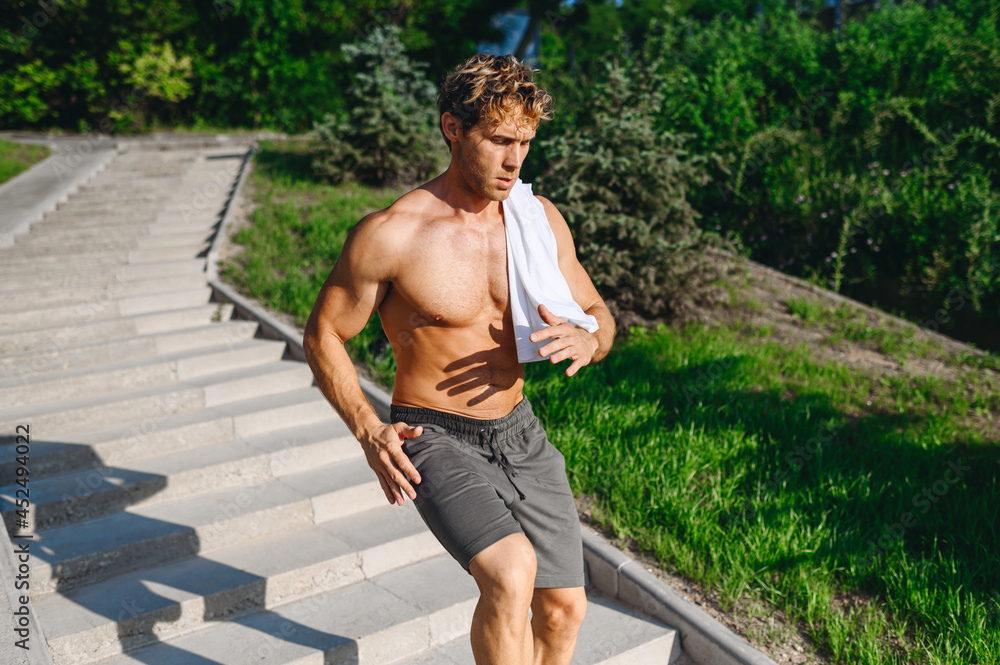 Young strong muscular sportsman caucasian man 20s with naked torso in shorts training walking jog run on steps up stairs in morning park on resort resting in leisure time. Helthy lifestyle concept