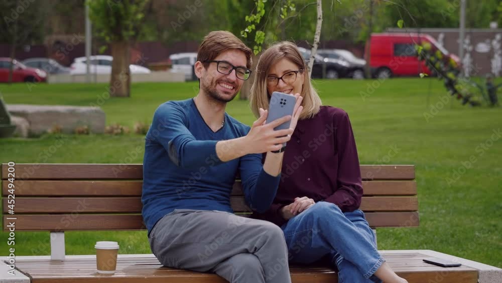 Happy couple wearing eye glasses waving hands at camera during video chat. Young cute man holds smartphone make video call. Video Call or watch video. Beautiful green grass birds background. Happy couple wearing eye glasses waving hands at camera during video chat. Young cute man holds smartphone make video call. Video Call or watch video. Beautiful green grass birds background.