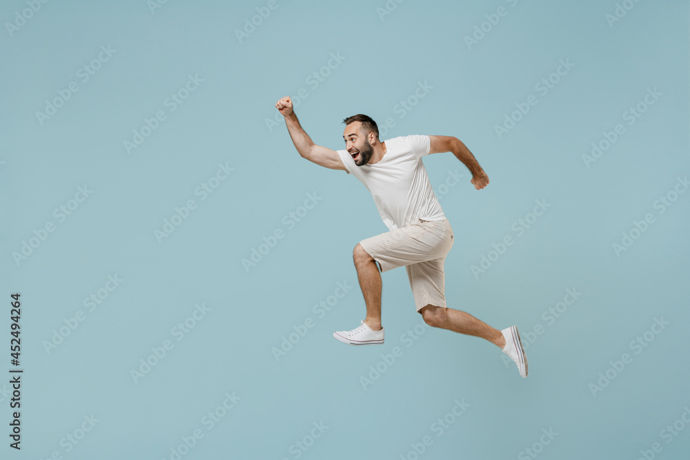Full length side view young excited happy sportsman runner man 20s wearing casual white t-shirt jump high run fast hurrying up isolated on plain pastel light blue color background studio portrait