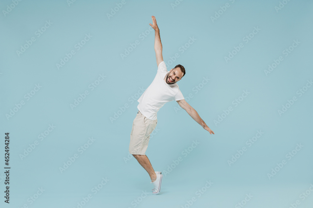 Full length young smiling happy man 20s in white t-shirt leaning back with outstretched hand dancing fooling around stand on toes isolated on plain pastel light blue color background studio portrait