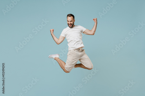 Full length young overjoyed excited happy caucasian man 20s wearing casual white t-shirt jump high do winner gesture clench fist isolated on plain pastel light blue color background studio portrait