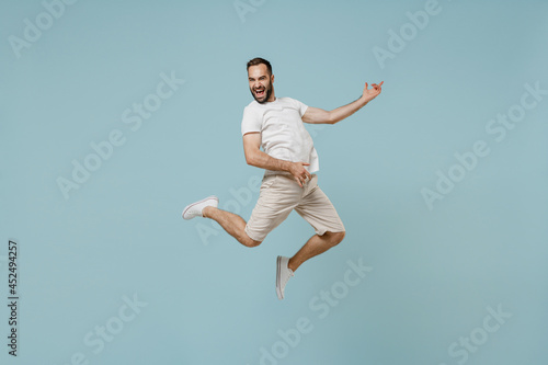 Full length young overjoyed expressive rock singer happy caucasian man 20s wearing casual white t-shirt jump high playing guitar isolated on plain pastel light blue color background studio portrait