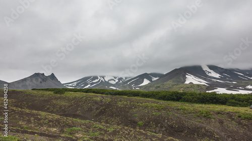 Dense gray clouds lie on the mountains. Areas of snow on the slopes. Sparse vegetation on the ground. A picturesque mountain against the background of a foggy sky in the distance. Kamchatka