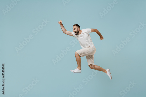 Full length side view young excited happy sportsman runner man 20s wearing casual white t-shirt jump high run fast hurrying up isolated on plain pastel light blue color background studio portrait