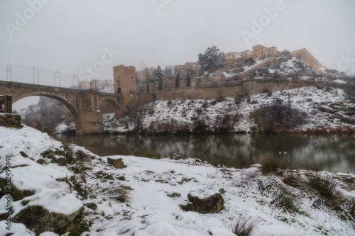 City landscape of snowy old city and castle with clouds and fog  View of the Tajo river and Alcántara bridge from Toledo during the storm Filomena, World Heritage, Spain. Horizontal view © Ricardo MzF .com 