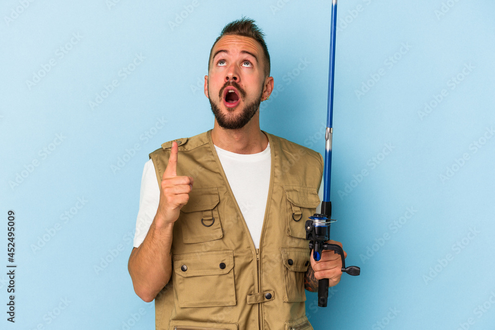 Young caucasian fisherman with tattoos holding rod isolated on blue background pointing upside with opened mouth. Young caucasian fisherman with tattoos holding rod isolated on blue background pointing upside with opened mouth.