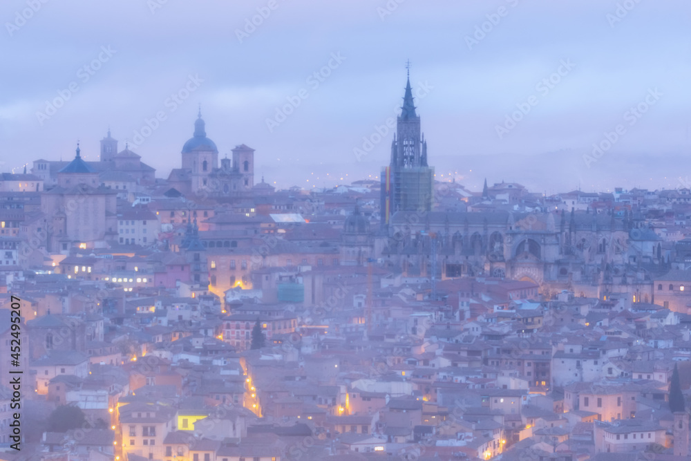 Cityscape in old city changing light at dawn; Tower of the primate cathedral with the old town and its small streets of Toledo with fog and clouds, world heritage site, Spain. Horizontal view