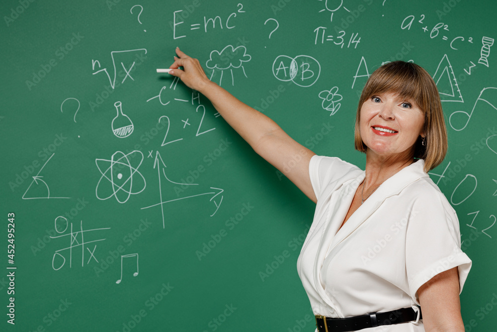 Attentive teacher mature elderly lady woman 55 wear white shirt hold in hand write point with chalk isolated on green wall blackboard background studio. Education in high school September 1 concept.