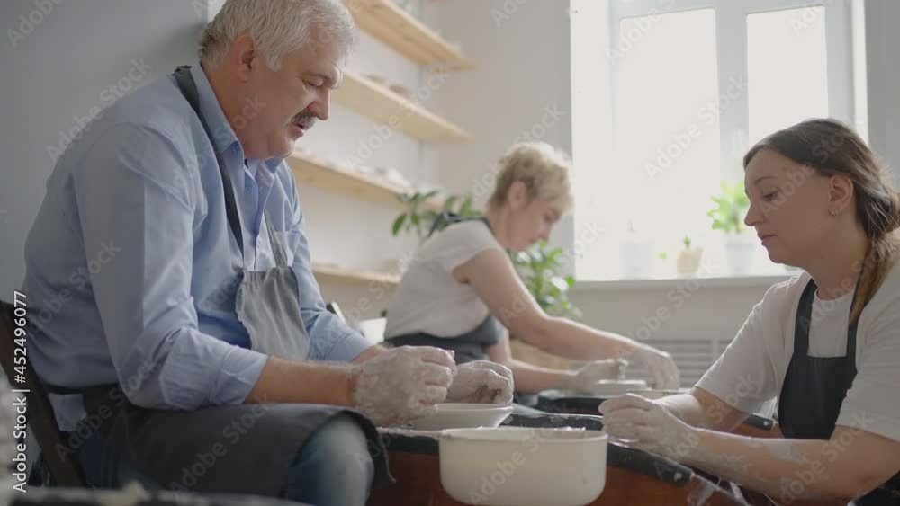 Medium shot of middle aged ceramic artist teaching group elderly Caucasian woman and senior man how to wedge clay sitting at desk in art studio. People enjoying talking at work Medium shot of middle aged ceramic artist teaching group elderly Caucasian woman and senior man how to wedge clay sitting at desk in art studio. People enjoying talking at work
