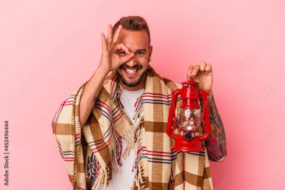 Young caucasian man with tattoos holding vintage lantern isolated on pink background  excited keeping ok gesture on eye.