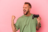 Young caucasian man with tattoos holding game controller isolated on pink background  raising fist after a victory, winner concept.