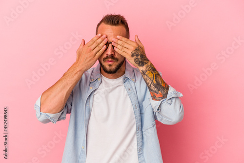 Young caucasian man with tattoos isolated on pink background  afraid covering eyes with hands.