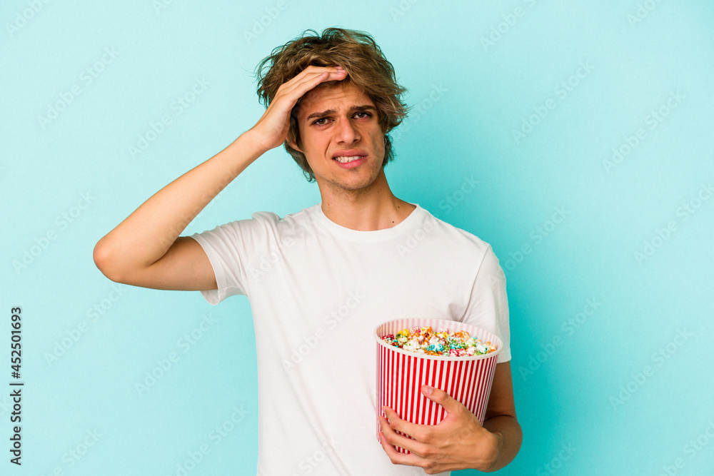 Young caucasian man with makeup holding popcorn isolated on blue background  being shocked, she has remembered important meeting.