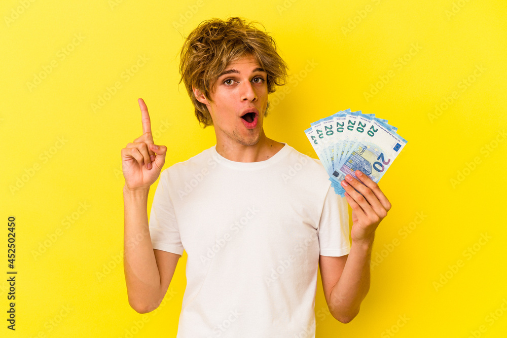 Young caucasian man with makeup holding bills isolated on yellow background having an idea, inspiration concept. Young caucasian man with makeup holding bills isolated on yellow background having an idea, inspiration concept.