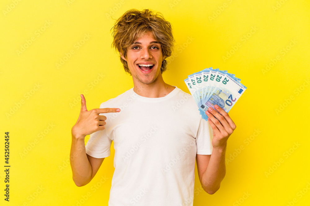 Young caucasian man with makeup holding bills isolated on yellow background person pointing by hand to a shirt copy space, proud and confident Young caucasian man with makeup holding bills isolated on yellow background person pointing by hand to a shirt copy space, proud and confident