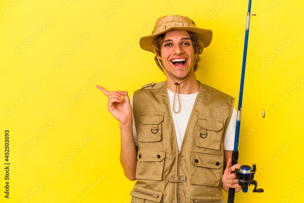 Young fisherman with makeup holding rod isolated on yellow background smiling and pointing aside, showing something at blank space. Young fisherman with makeup holding rod isolated on yellow background smiling and pointing aside, showing something at blank space.