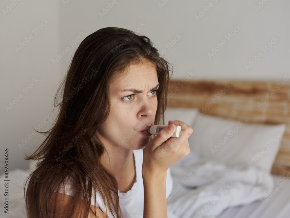 woman holding a thermometer in her mouth while sitting on the bed temperature health problems woman holding a thermometer in her mouth while sitting on the bed temperature health problems