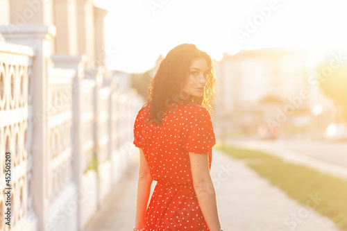 Beautiful young caucasian woman with curly brunette hair in a retro bright red dress walks in the city at sunset