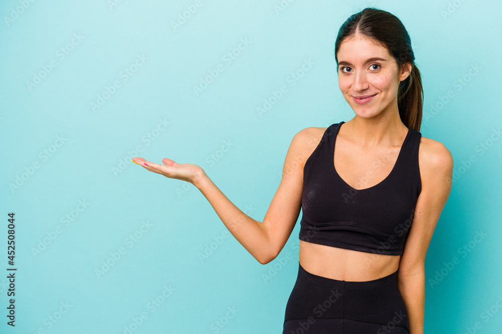 Young sporty caucasian woman isolated on blue background showing a copy space on a palm and holding another hand on waist. Young sporty caucasian woman isolated on blue background showing a copy space on a palm and holding another hand on waist.