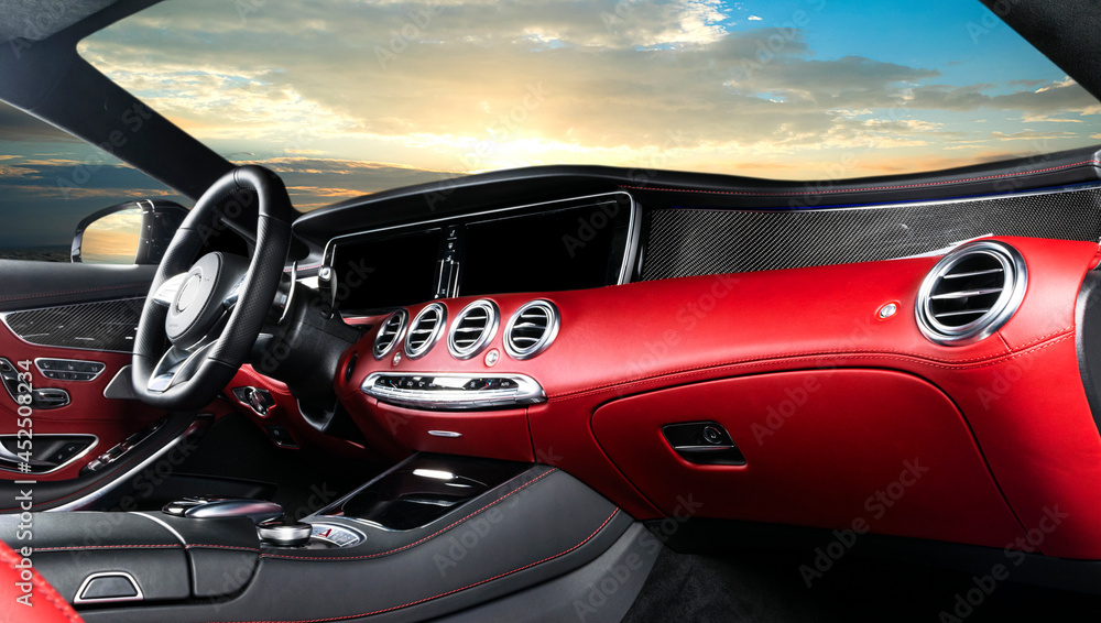 Red luxury modern car Interior. Steering wheel and dashboard. Detail of modern car interior. Automatic gear stick. Leather seats with stitching in expensive car. Sunset sky background