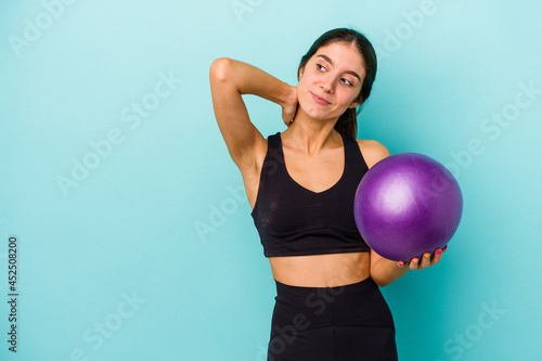 Young caucasian fitness woman holding a ball isolated on blue background touching back of head, thinking and making a choice.