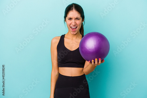 Young caucasian fitness woman holding a ball isolated on blue background screaming very angry and aggressive.