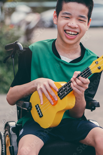 Happy handicapped teenager boy on wheelchair play yellow ukulele with smiling face, Lifestyle of disabled child with music therapy is good for mental health.