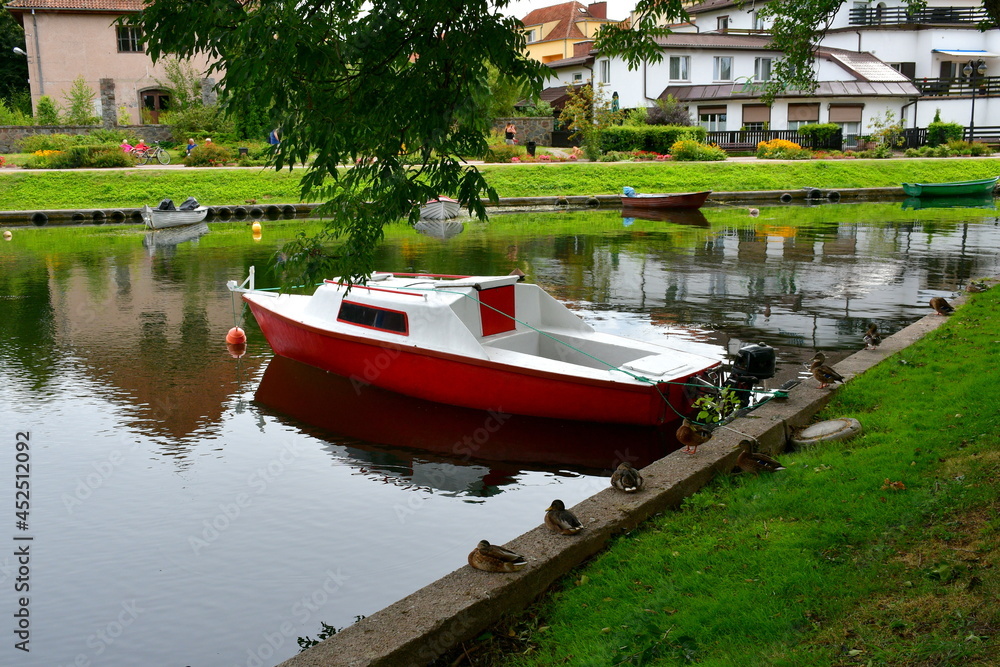 Obraz premium Close up on some ducks standing and sitting on a concrete edge or a river or lake located next to the spot where various passenger boats and other vessels are parked next to a holiday resort in Poland