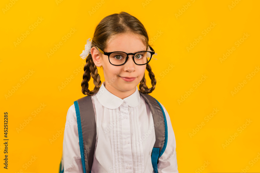 Back to school. Girl kid 7 y.o. with pigtails made of hair, glasses and with a backpack looking at camera in isolation on a yellow background.